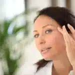 A woman touches her face while looking in a mirror, assessing the effects of her eyelid surgery