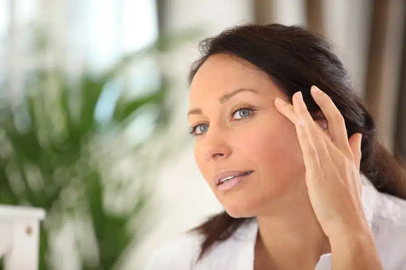 A woman touches her face while looking in a mirror, assessing the effects of her eyelid surgery